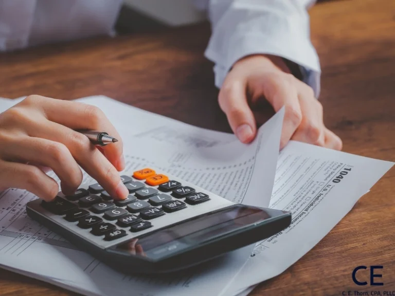 A person's hand typing on a calculator with a pen and 1040. This image accompanies the post titled The Pros and Cons of Making Quarterly Estimated Tax Payments.