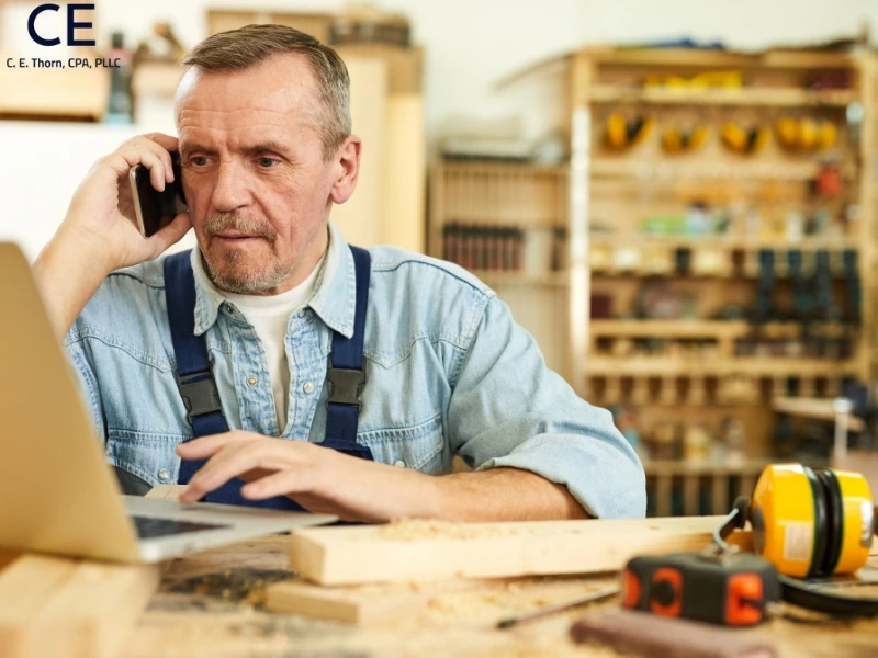 A man on a laptop in a workshop asks his account "can you write off tax prep fees?" on the phone.