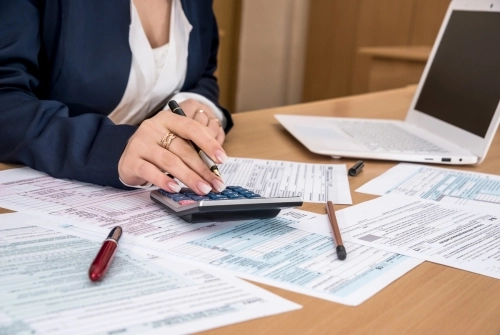 A woman CPA uses a calculator and has tax forms on her desk as she files business taxes and avoids common tax mistakes.